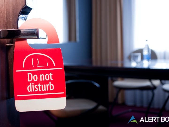 AlertBot blog titled "Why Your Website Monitoring Solution Needs a Do-Not-Disturb Feature" with a photo of a hotel room out of focus in the background and a "Do Not Disturb" sign on the doorknob in the foreground. AlertBot logo is placed in the lower right.